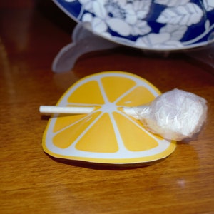 Op de afbeelding: Een witte lolly met een gele, citroen-vormige papieren verpakking. De lolly ligt op een houten oppervlak.