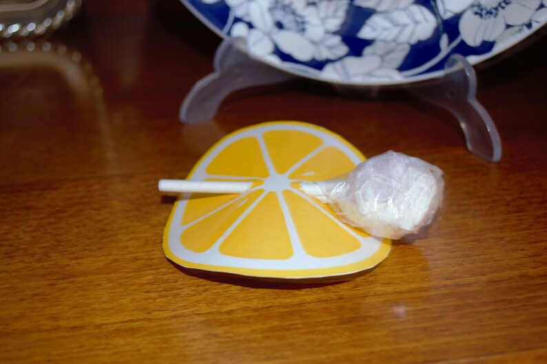 Op de afbeelding: Een witte lolly met een gele, citroen-vormige papieren verpakking. De lolly ligt op een houten oppervlak.