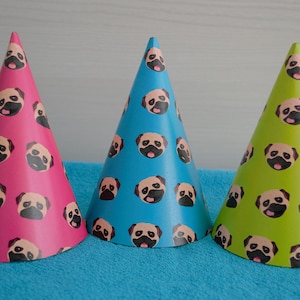 Puede incluir: Tres sombreros de fiesta con un patrón de perro pug. Los sombreros son rosa, azul y verde.
