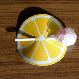Op de afbeelding: Een gele citroenschijf van papier met een witte stok en een roze en witte lolly die in het midden is bevestigd.