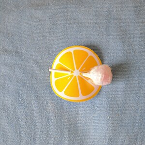 Op de afbeelding: Een roze lolly met een witte stok is geplaatst op een gele citroenschijf-uitsparing. De citroenschijf-uitsparing is op een blauw, getextureerd oppervlak.