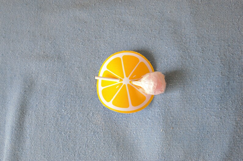 Op de afbeelding: Een roze lolly met een witte stok is geplaatst op een gele citroenschijf-uitsparing. De citroenschijf-uitsparing is op een blauw, getextureerd oppervlak.