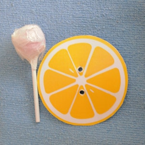 Op de afbeelding: Een roze lolly met een witte stok op een blauwe achtergrond naast een gele citroenschijf met witte lijnen.