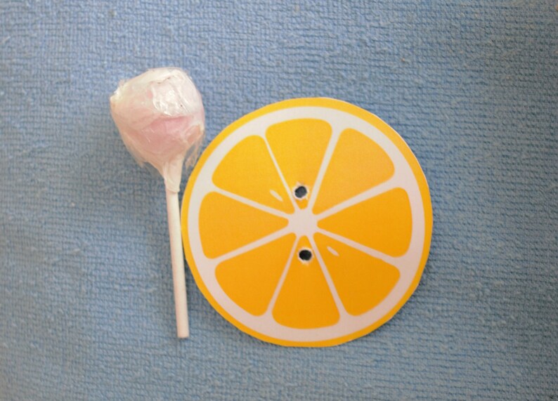 Op de afbeelding: Een roze lolly met een witte stok op een blauwe achtergrond naast een gele citroenschijf met witte lijnen.