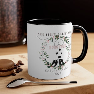 May include: White ceramic mug with a black rim and handle. The mug features a wreath with birds, hearts, and the text "Our First Christmas Together" and "Emily & Alfred 2022".