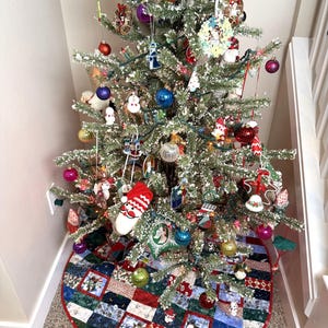 May include: A decorated Christmas tree with various ornaments, including colorful baubles, snowmen, and Santa-themed decorations. The tree is placed in a corner and sits on a patchwork tree skirt with penguin and floral patterns.