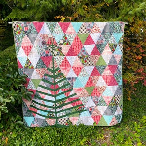 May include: A colorful quilt with a Christmas tree design. The quilt features a patchwork of triangles in red, green, white, and blue fabrics. A green tree is appliqued on the front, with a star-like embellishment at the top. The quilt is displayed outdoors.