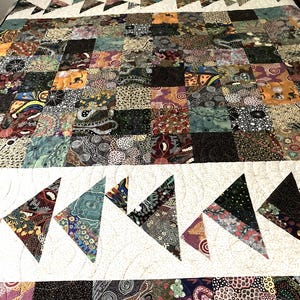 May include: A patchwork quilt with a variety of patterned squares in shades of brown, green, and purple. White bands with triangle designs separate the sections. The quilt is on a bed.