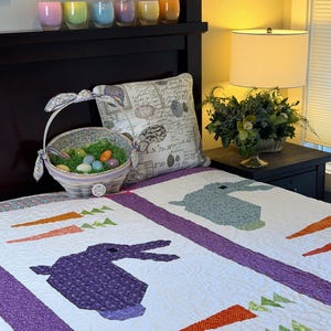 May include: A white quilt with purple borders and appliqued bunnies and carrots. A decorative Easter basket with eggs sits on the bed. Six colorful glass candle holders are on a shelf above the bed.