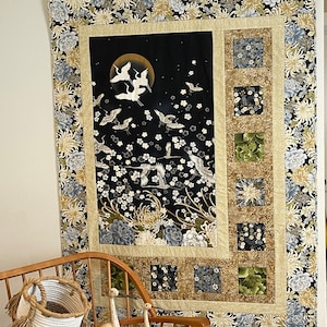 May include: A black and gold wall hanging with a floral pattern and white cranes flying in front of a full moon. The wall hanging is framed with a gold border and a floral pattern.