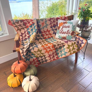 May include: A patchwork quilt featuring a repeating star pattern in shades of orange, teal, and brown, draped over a wooden bench. A white throw pillow with the text "give thanks" and a pumpkin and leaf design sits on the quilt near four decorative pumpkins.