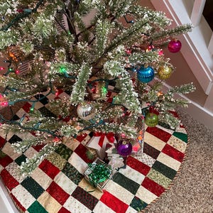 May include: A decorated Christmas tree with colorful ornaments and lights, set on a quilted tree skirt. The skirt features a patchwork design in red, green, and cream squares. Presents are placed under the tree, adding to the festive holiday scene.