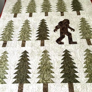 May include: A quilt featuring a repeating pattern of green pine trees on a light cream background. One dark brown silhouette of a Bigfoot figure is integrated into the grid of trees. The quilt has a red border along the top edge.