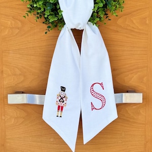 Puede incluir: Una corona de puerta de tela blanca con un cascanueces y la letra "S" bordada. La corona está colgada en una puerta de madera con una guirnalda de hojas verdes.