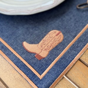 May include: A blue denim placemat with a brown embroidered cowboy boot design. The placemat has a brown stitched border.