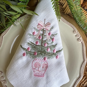 May include: White linen napkin featuring an embroidered Christmas tree design. The tree is green with pink baubles and a pink bow. The tree sits in a pink vase. The napkin is folded and placed on a cream-coloured plate.
