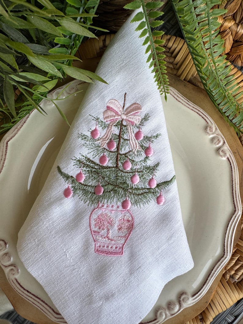 May include: White linen napkin featuring an embroidered Christmas tree design. The tree is green with pink baubles and a pink bow. The tree sits in a pink vase. The napkin is folded and placed on a cream-coloured plate.