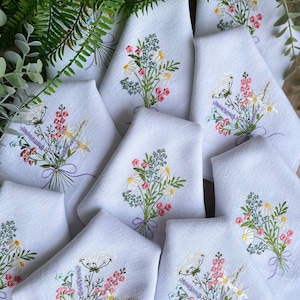 May include: A set of 10 white linen napkins featuring embroidered floral bouquets in shades of pink, white, and green. The bouquets are tied with a purple ribbon.