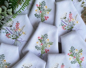 Puede incluir: Un juego de 10 servilletas de lino blanco con bordados de ramos florales en tonos rosa, blanco y verde. Los ramos están atados con una cinta morada.