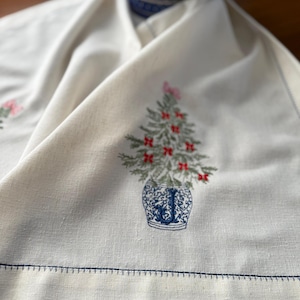 May include: Off-white linen Christmas table runner with a blue stitched border. The runner features an embroidered Christmas tree in a blue and white vase with red bows. A blue and white patterned bowl is in the background.