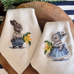 May include: Two embroidered linen tea towels featuring a bunny design. One bunny is wearing blue overalls and holding a bouquet of yellow flowers. The other bunny is wearing a blue dress and holding a bouquet of yellow flowers.
