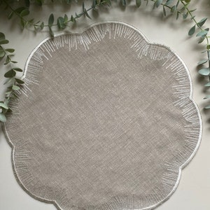 Puede incluir: Un mantel individual de lino beige con borde festoneado y detalles bordados en blanco.