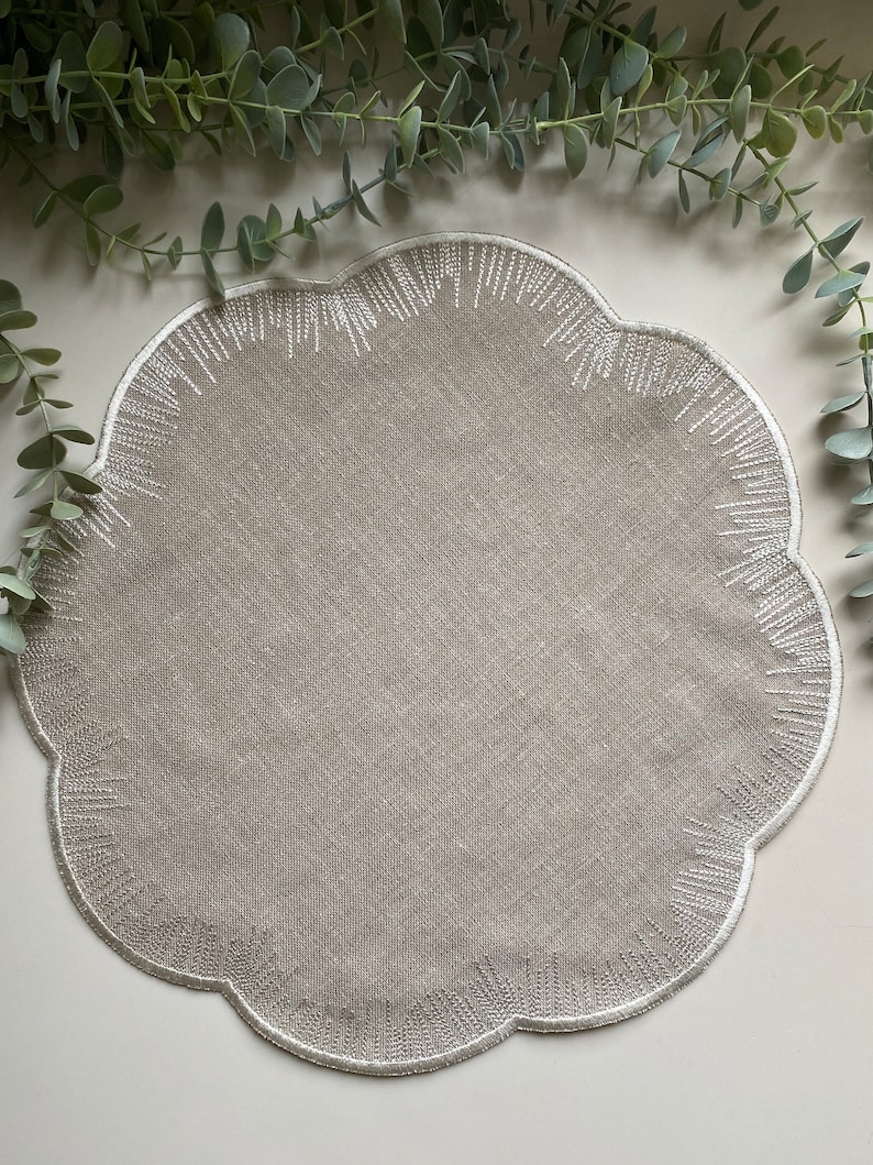 Puede incluir: Un mantel individual de lino beige con borde festoneado y detalles bordados en blanco.