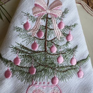 May include: White napkin with an embroidered Christmas tree design. The tree is decorated with pink baubles and a pink bow. The tree is in a pink vase. The napkin is folded into a triangle.