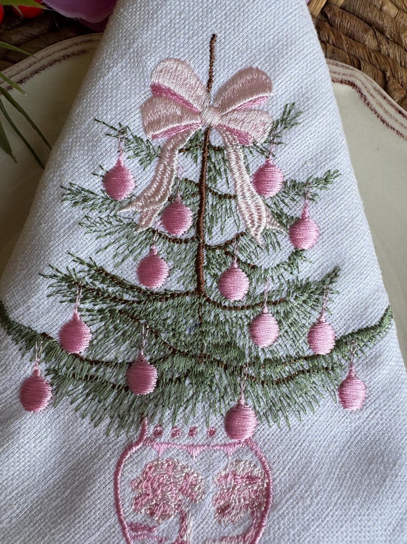 May include: White napkin with an embroidered Christmas tree design. The tree is decorated with pink baubles and a pink bow. The tree is in a pink vase. The napkin is folded into a triangle.