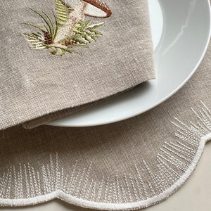Puede incluir: Un mantel individual de lino beige con borde festoneado y detalles bordados en blanco. Un plato blanco con una servilleta de lino beige con un dise&ntilde;o de hongo bordado se coloca sobre el mantel individual.