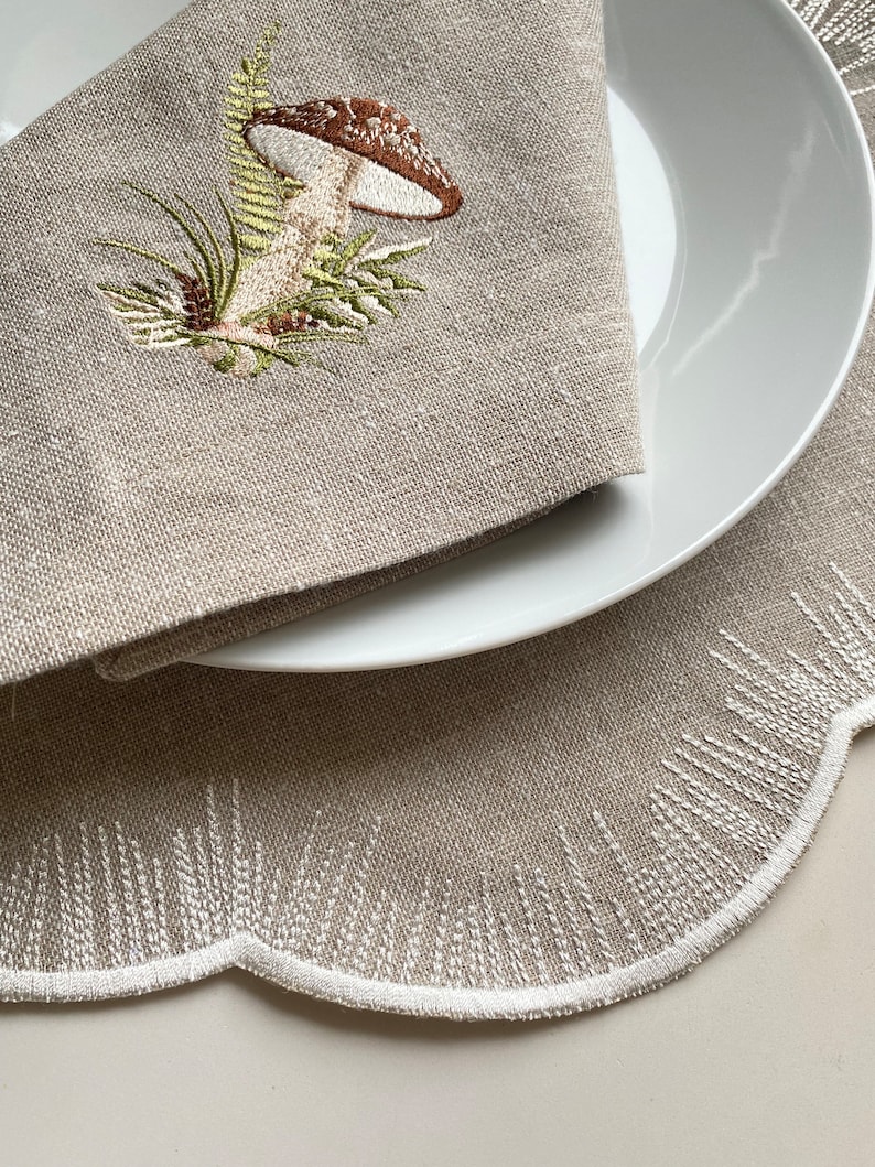 Puede incluir: Un mantel individual de lino beige con borde festoneado y detalles bordados en blanco. Un plato blanco con una servilleta de lino beige con un dise&ntilde;o de hongo bordado se coloca sobre el mantel individual.