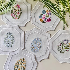 Puede incluir: Conjunto de seis posavasos de lino blanco con diseños florales bordados en varios colores. Cada posavasos presenta un diseño floral diferente en forma de huevo.