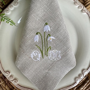 May include: A beige linen napkin with embroidered white snowdrops and green stems. The napkin is folded and placed on a white plate with a brown wicker placemat.