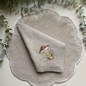 Puede incluir: Un mantel individual de lino beige con borde festoneado y bordado blanco. Una servilleta de lino doblada con un dise&ntilde;o de hongo bordado est&aacute; encima del mantel individual.