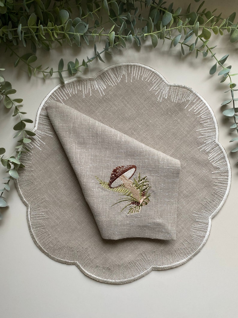 Puede incluir: Un mantel individual de lino beige con borde festoneado y bordado blanco. Una servilleta de lino doblada con un dise&ntilde;o de hongo bordado est&aacute; encima del mantel individual.