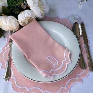May include: A pink linen napkin with a scalloped edge and a white embroidered letter "D" on it. The napkin is placed on a white plate with a pink scalloped charger underneath.