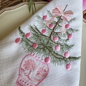 May include: White linen napkin featuring an embroidered Christmas tree design. The tree is green with pink baubles and a pink bow. The tree sits in a pink vase. The napkin is folded into a triangle.