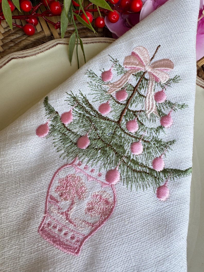May include: White linen napkin featuring an embroidered Christmas tree design. The tree is green with pink baubles and a pink bow. The tree sits in a pink vase. The napkin is folded into a triangle.