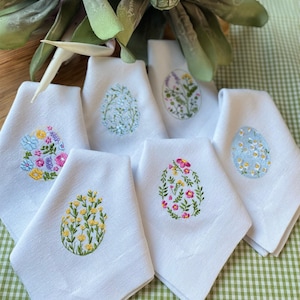 Pode incluir: Guardanapos de linho branco com bordados de ovos de Páscoa. Cada guardanapo apresenta um padrão floral único em várias cores, incluindo amarelo, rosa, azul e verde. Os guardanapos estão dispostos sobre uma toalha de mesa xadrez verde e branca.
