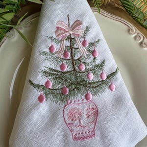May include: White linen napkin featuring an embroidered Christmas tree design. The tree is green with pink baubles and a pink bow. The tree sits in a pink vase. The napkin is folded on a white plate.