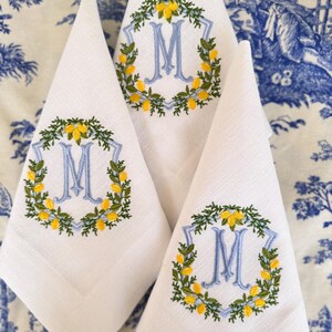 Puede incluir: Tres servilletas de lino blanco con guirnaldas de limón bordadas y la letra "M" en azul. Las guirnaldas son verdes con limones amarillos.