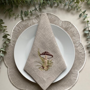 Puede incluir: Una servilleta de lino beige con un dise&ntilde;o de hongo bordado. La servilleta est&aacute; doblada y colocada sobre un plato blanco, que est&aacute; encima de un mantel individual beige con borde festoneado.