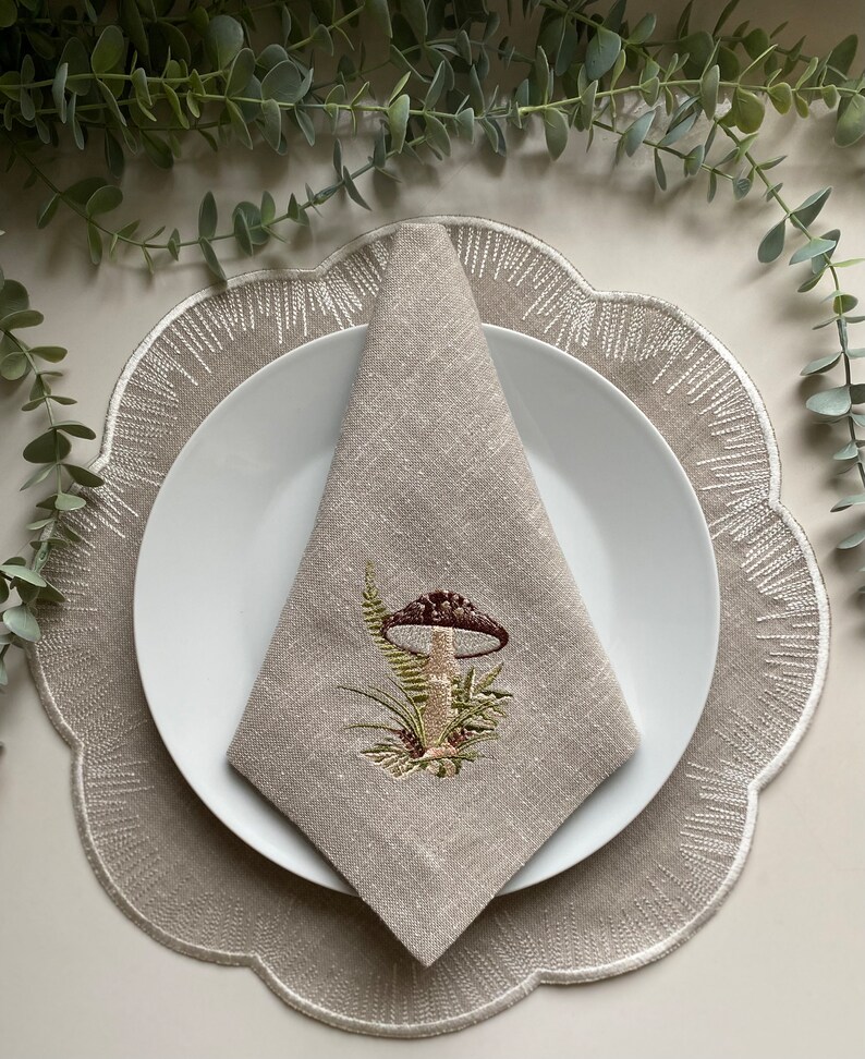 Puede incluir: Una servilleta de lino beige con un dise&ntilde;o de hongo bordado. La servilleta est&aacute; doblada y colocada sobre un plato blanco, que est&aacute; encima de un mantel individual beige con borde festoneado.