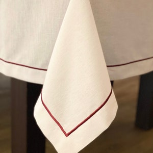 May include: White linen tablecloth with a burgundy red border. The tablecloth is folded in half and draped over a table.
