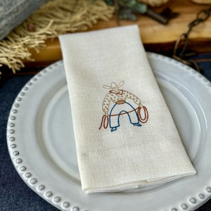 Puede incluir: Una servilleta de lino blanco con un diseño bordado en marrón, azul y rojo de un vaquero sosteniendo un lazo. La servilleta está doblada y colocada en un plato blanco con un borde de cuentas.