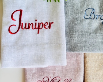 Puede incluir: Primer plano de cuatro toallas de mano personalizadas en blanco, gris, rosa y beige. La toalla blanca tiene "Juniper" bordado en rojo. La toalla gris tiene "Bro" bordado en azul. La toalla rosa tiene "Molly" bordado en burdeos.