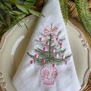 May include: White linen napkin featuring an embroidered Christmas tree. The tree is green with pink baubles and a pink bow. The tree is in a pink vase. The napkin is folded on a cream plate with a decorative edge.
