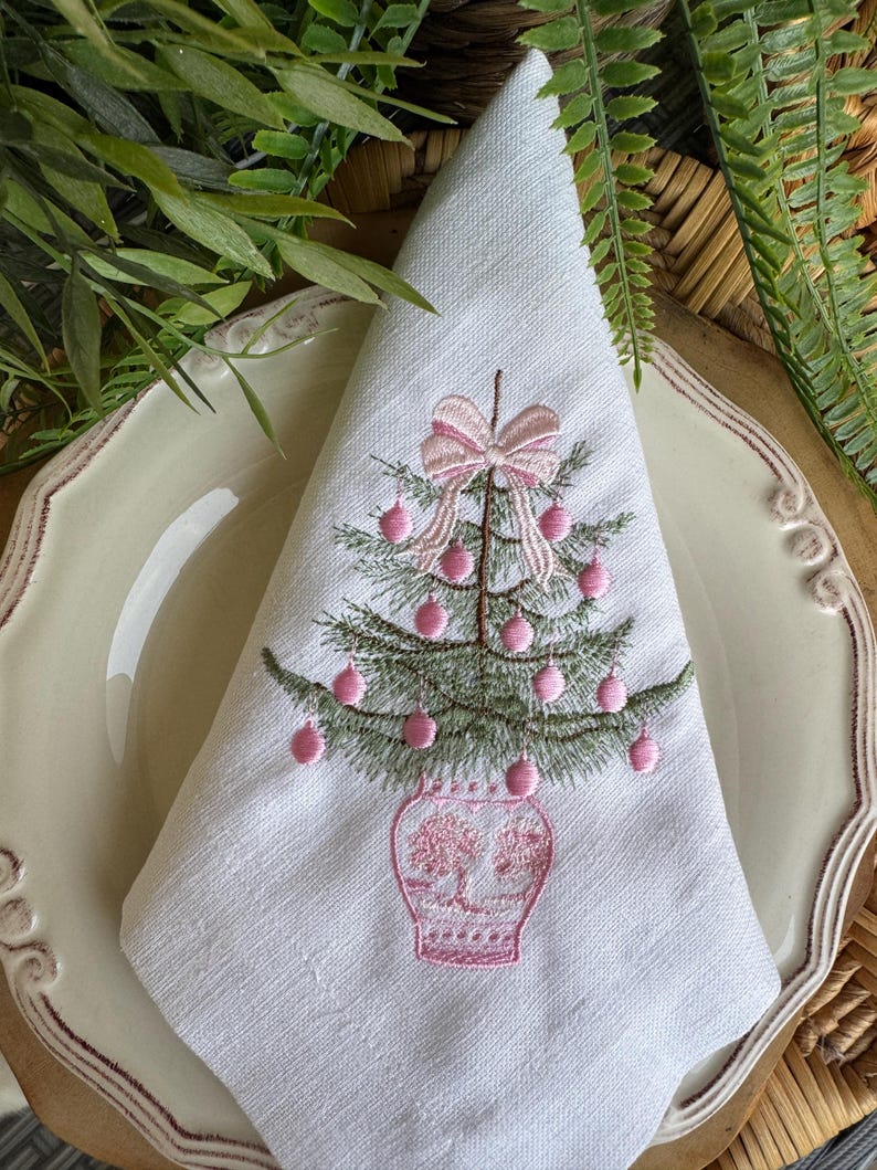 May include: White linen napkin featuring an embroidered Christmas tree. The tree is green with pink baubles and a pink bow. The tree is in a pink vase. The napkin is folded on a cream plate with a decorative edge.