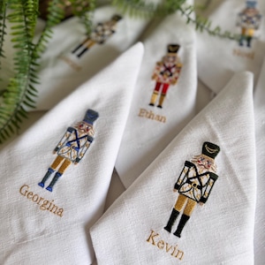 Op de afbeelding: Witte stoffen servetten geborduurd met kleurrijke notenkraker soldaten ontwerpen. Elke servet heeft een andere notenkraker en een naam geborduurd in gouden draad, waaronder "Georgina" en "Kevin". De servetten zijn gevouwen en gerangschikt op een wit oppervlak.