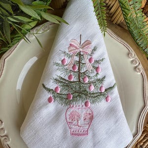 May include: White linen napkin featuring an embroidered Christmas tree design. The tree is green with pink baubles and a pink bow. The tree sits in a pink vase. The napkin is folded and placed on a white plate with a decorative edge.